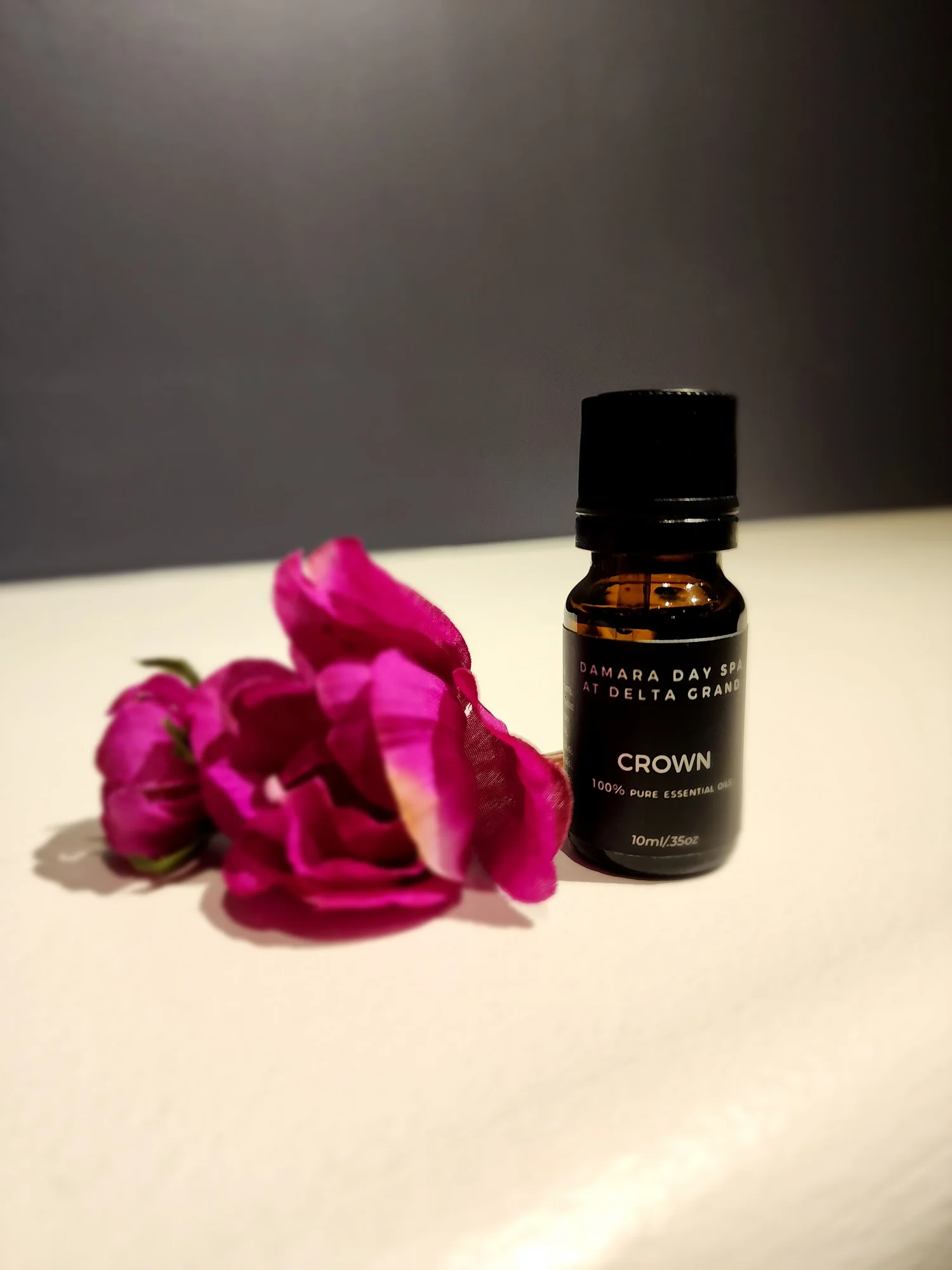 20231123_152125 – Damara Luxury Spa Vancouver Brown glass essential oil bottle with a black cap next to a purple-pink flower on a light surface. The label hints at spa use and natural oils.