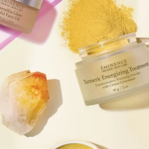 Flat lay of Eminence skincare products: a jar of Turmeric Energizing Treatment, a small Glow Oil jar, a pile of turmeric powder, a crystal, and an open yellow cream jar on a pale pink background with a diagonal pink line.