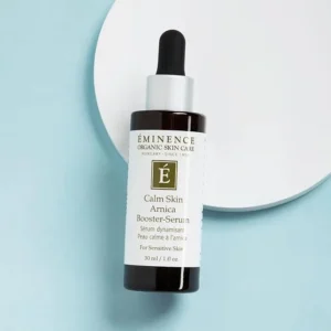 Bottle of Eminence Calm Skin Arnica Booster-Serum with dropper on a pastel blue background on a white circular surface behind it
