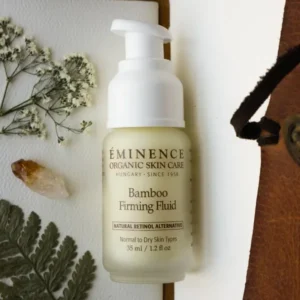 Bottle of Eminence Bamboo Firming Fluid with pump, placed on a white surface with dried flowers and ferns nearby.
