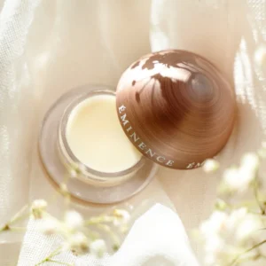 Open jar of Eminence skincare cream resting on white fabric, with a brown swirl-cap and delicate white flowers nearby.