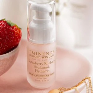 Eminence Strawberry Rhubarb Hyaluronic Hydrator bottle with pump on a pink surface, with a strawberry and gold jewelry nearby (soft aesthetic).
