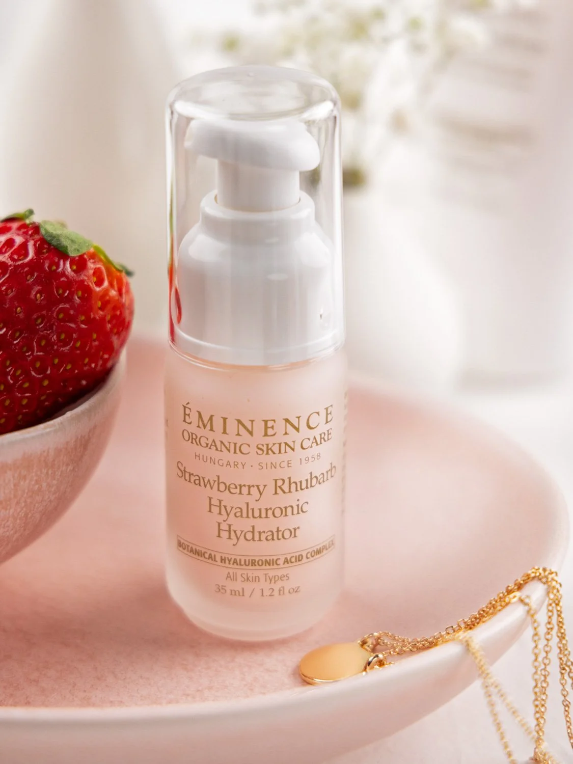 EM164-1164-strawberry-rhubarb-hyaluronic-hydrator-pdp-library-image-1125x1500 – Damara Luxury Spa Vancouver Eminence Strawberry Rhubarb Hyaluronic Hydrator bottle with pump on a pink surface, with a strawberry and gold jewelry nearby (soft aesthetic).