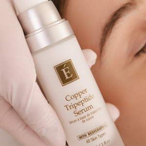 Close-up of a Copper Tripeptide Serum bottle being held by a gloved hand next to a person's face, highlighting the product.