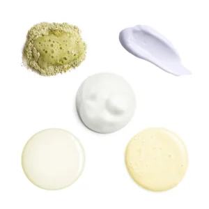 Flat lay of skincare products: dusting powder with a paste, a lilac cream smear, white foamy lotion, and two pale round creams on white background