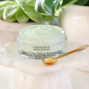 Jar of Eminence Stone Crop Whip Moisturizer on a marble stand with a wooden spoon and green plants in the background, skincare product context.