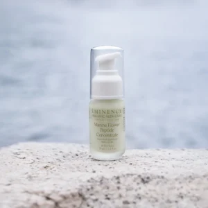 A skincare serum bottle (Marine Flower Peptide Concentrate) on a textured rock with the ocean blurred in the background.