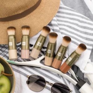 Flat lay of makeup brushes lined beside a beige straw hat on striped fabric, with sunglasses and a bowl of green apples nearby.