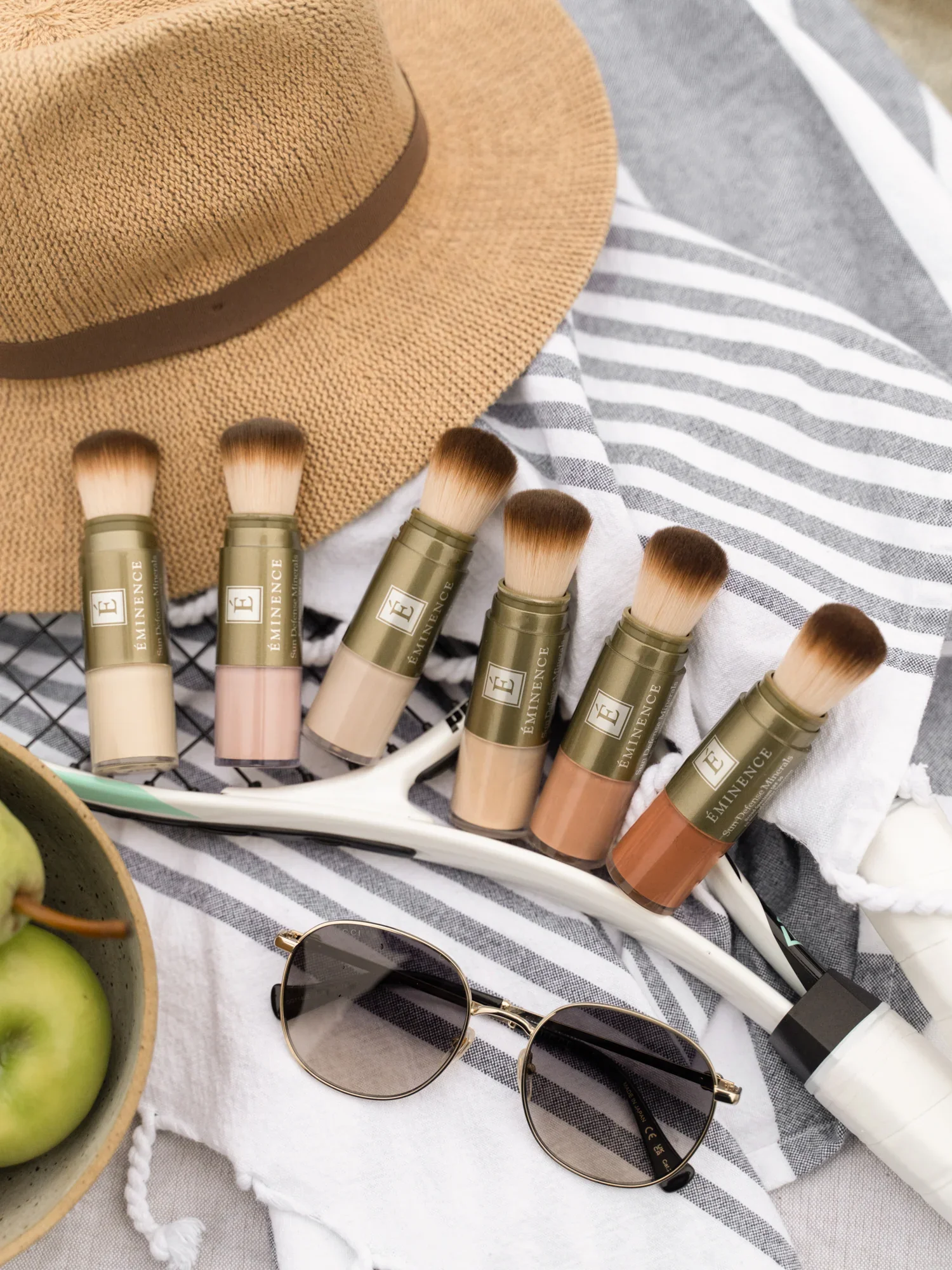 Spring-2025-Launch-Lifestyle-1080x1350-09 – Damara Luxury Spa Vancouver Flat lay of makeup brushes lined beside a beige straw hat on striped fabric, with sunglasses and a bowl of green apples nearby.