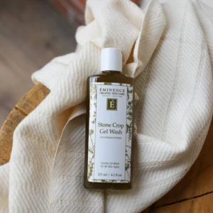 Bottle of Éminence Stone Crop Gel Wash resting in a soft cream towel on a wooden surface; front label visible.