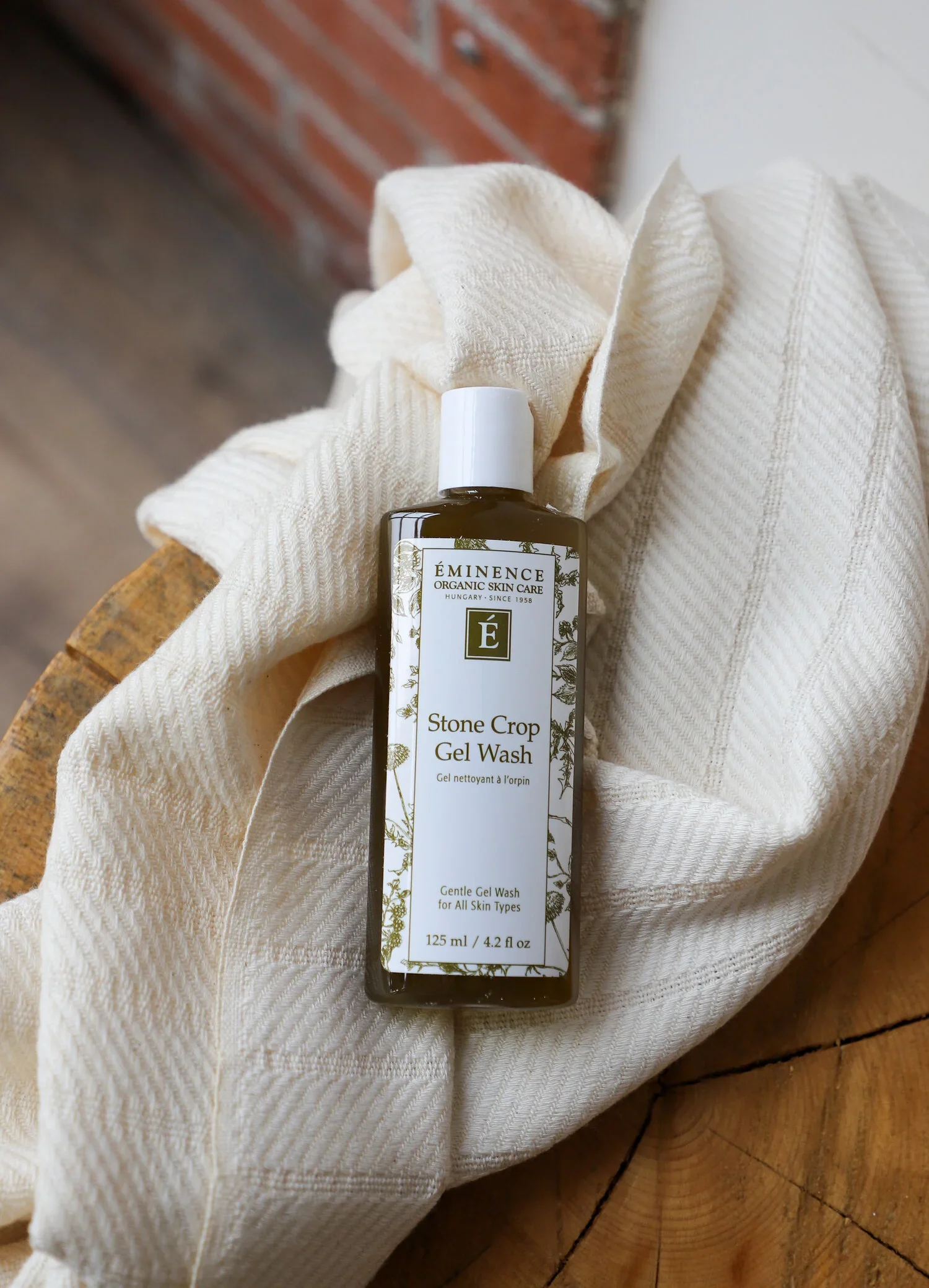 Stone_Crop_Gel_Wash_2 – Damara Luxury Spa Vancouver Bottle of Éminence Stone Crop Gel Wash resting in a soft cream towel on a wooden surface; front label visible.