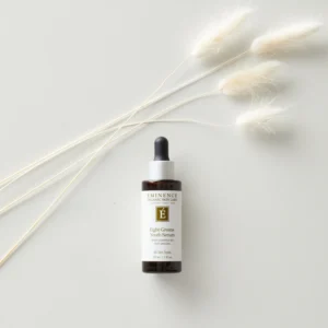 Dropper bottle of Eminence Eight Greens Youth Serum on a pale surface with wispy dried grasses nearby guiding the eye to the product.