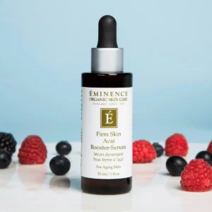 Bottle of Eminence Firm Skin Acai Booster-Serum with dropper, surrounded by raspberries and blueberries on a light surface.